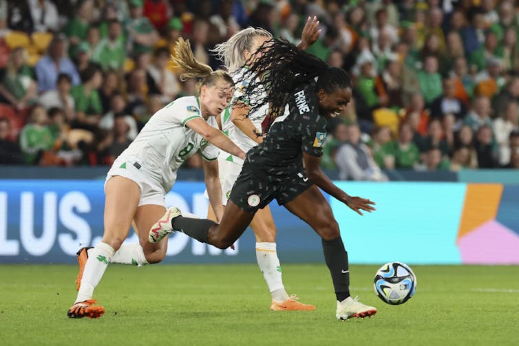 A Black women in a black soccer uniform dribbles a soccer ball away from two white women in white soccer uniforms