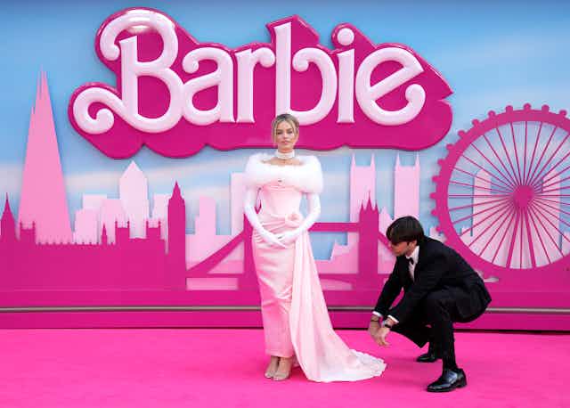 A woman in a light pink gown, with Barbie in large letters written behind her, poses on a pink carpet.