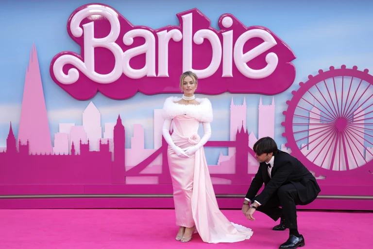 A woman in a light pink gown, with Barbie in large letters written behind her, poses on a pink carpet.