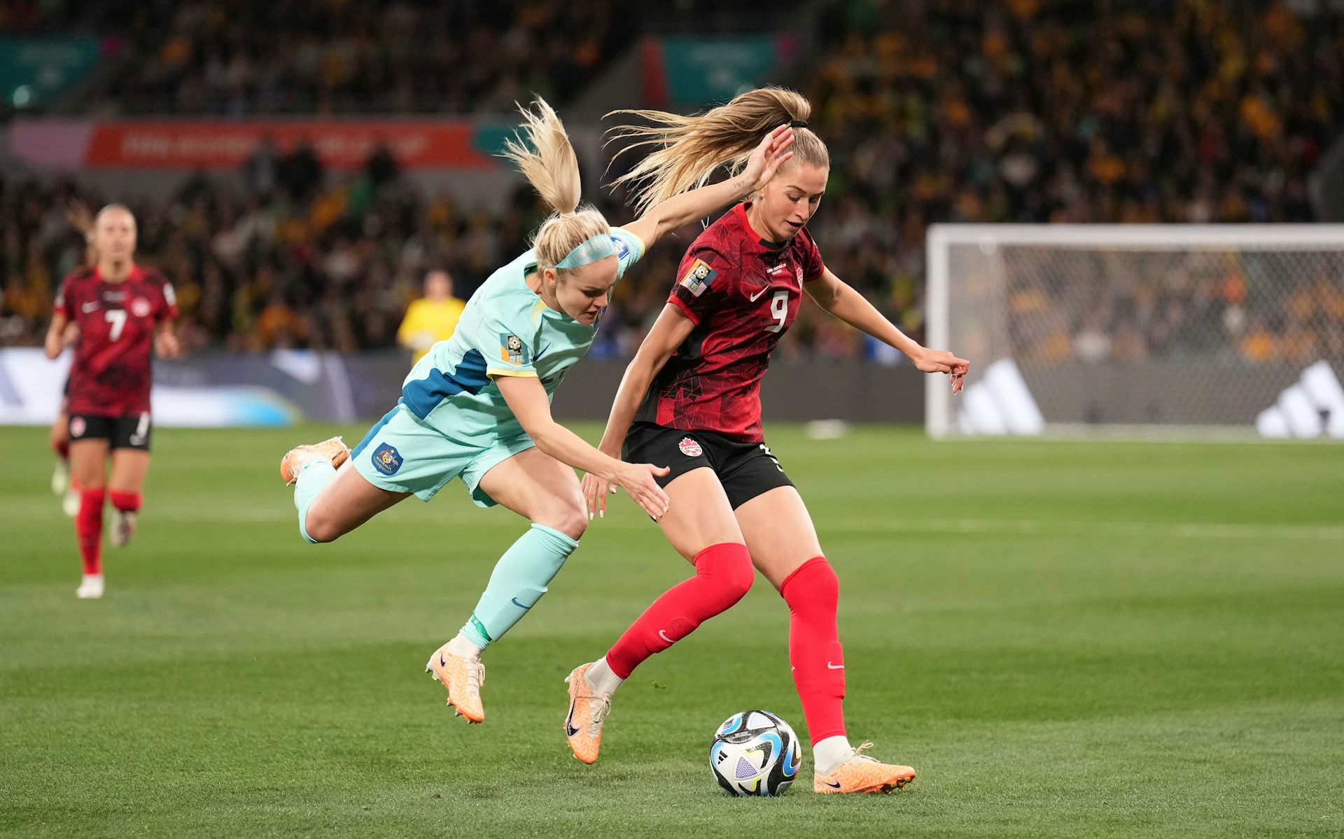 Dos futbolistas rubias luchan por un balón, una vestida de azul claro y saltando hacia el hombro izquierdo de otra vestida de rojo y negro