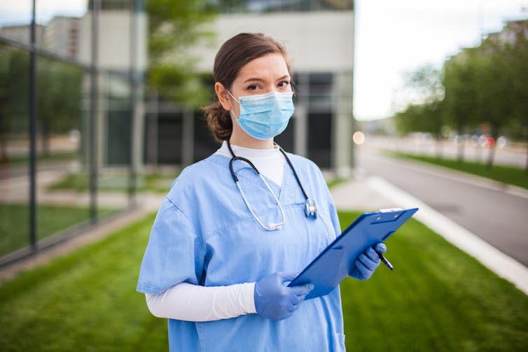 Health worker wearing surgical mask, scrubs and gloves outside carrying folder or clipboard