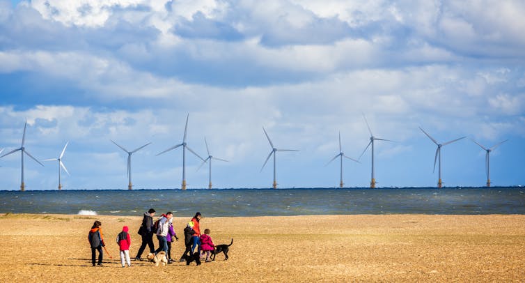 Uma família passeando com cães em uma praia em frente a um parque eólico offshore.