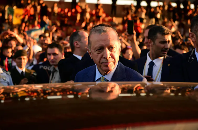 Turkish president Recep Tayyip Erdoğan about to get into a car with his bodyguards and a crowd of supporters in the background.