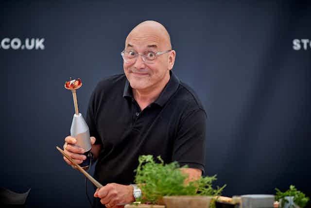 Man with glasses and black shirt holding kitchen appliance in front of a black screen, herb plants in the foreground.