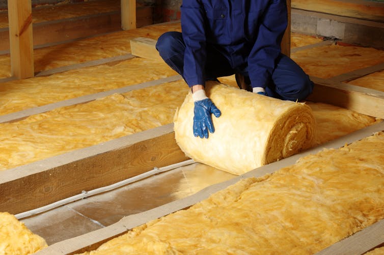 A worker in blue overalls rolls out yellow foam cladding in an attic.