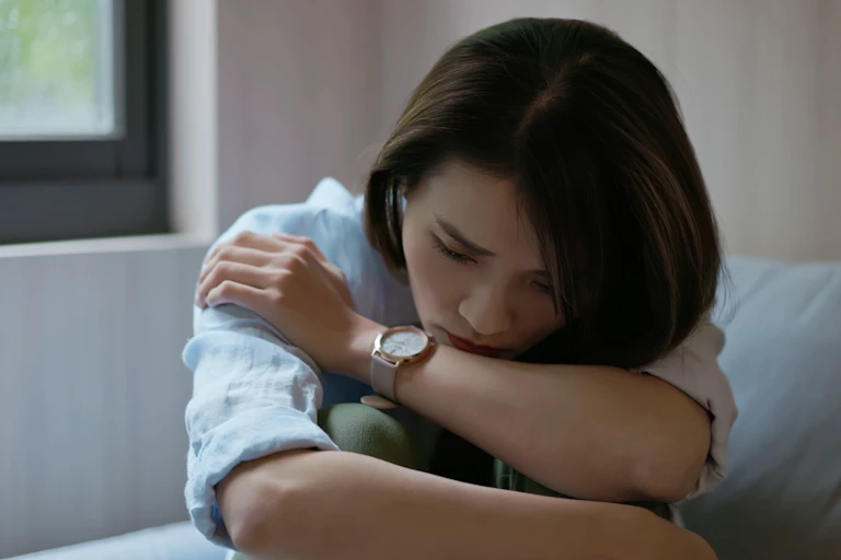 A young Chinese woman with a sad expression on her face, sitting on a couch and holding her knees to her chest
