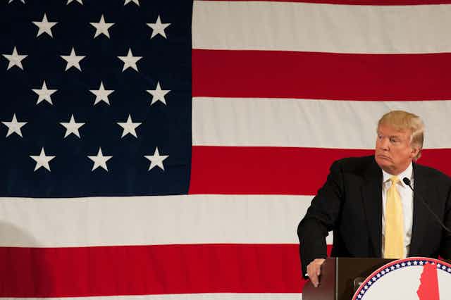 El expresidente Donald Trump tras un atril mira de lado con la bandera estadounidense de fondo.