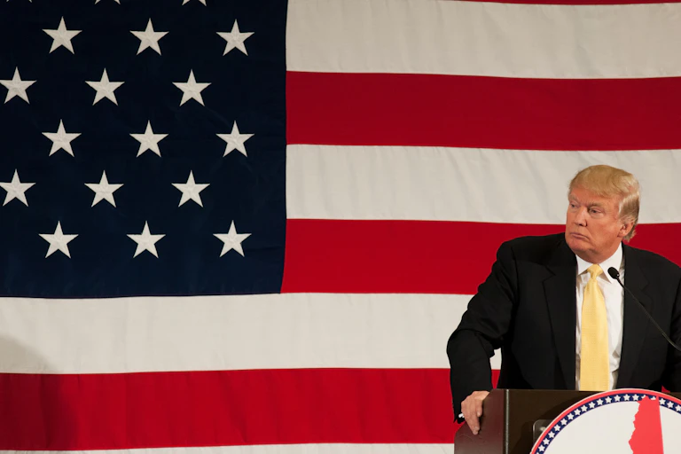 El expresidente Donald Trump tras un atril mira de lado con la bandera estadounidense de fondo.
