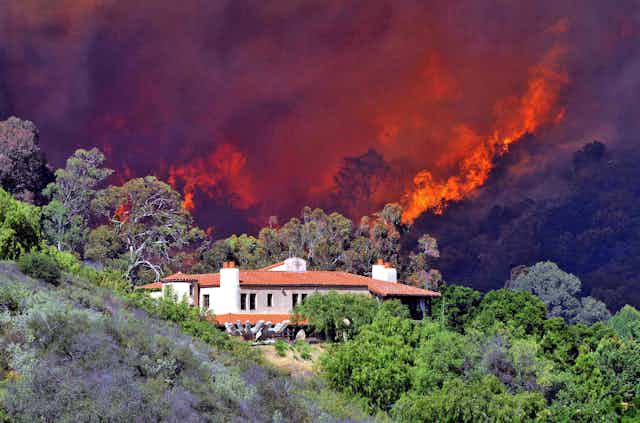 Fire rages in the hills behind a home surrounded by dry bushes and trees.