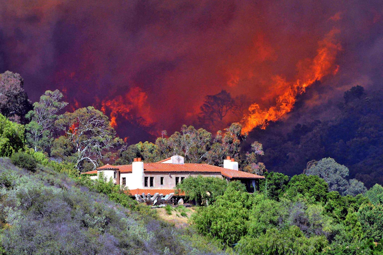 Fire rages in the hills behind a home surrounded by dry bushes and trees.