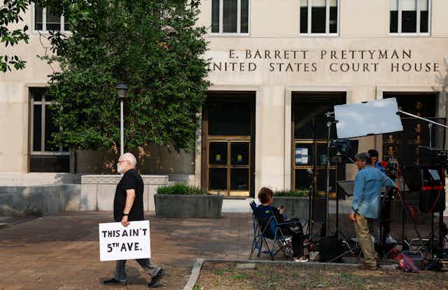 A man walks by a courthouse and holds a sign that says 'This ain't 5th ave,' next to a group of people who appear tone photographers and reporters.
