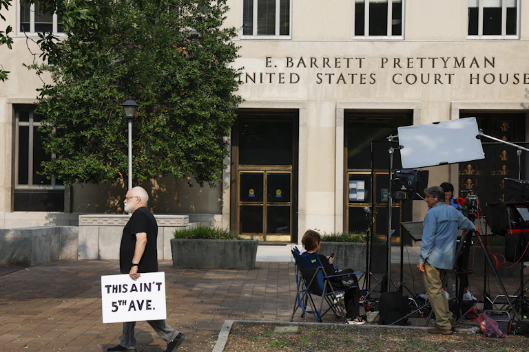 A man walks by a courthouse and holds a sign that says 'This ain't 5th ave,' next to a group of people who appear tone photographers and reporters.