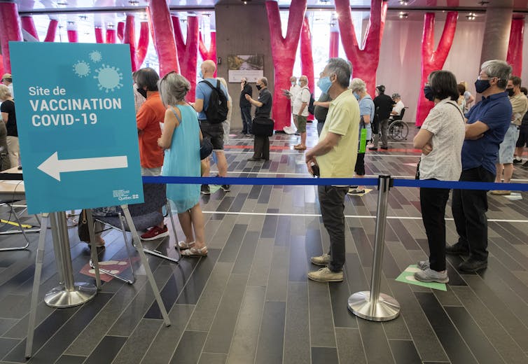 People in a socially distanced queue behind a blue sign with an arrow to a vaccine clinic