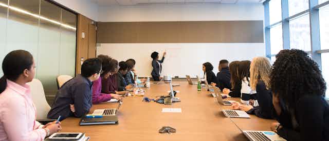 a Black woman stands at a whiteboard and leads a workshop to a diverse group at a big table