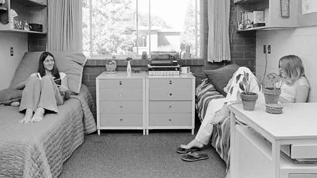Two students seen in a dorm room, each sitting on their respective beds.