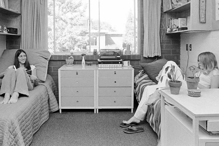 Two students seen in a dorm room, each sitting on their respective beds.