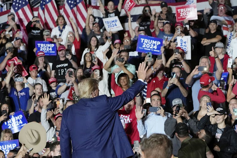A man is seen from behind waving to supporters waving Trump 2024 signs.