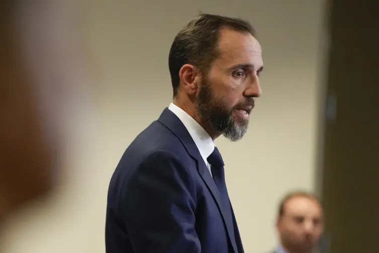 A dark-haired bearded man in a dark suit at a news conference.