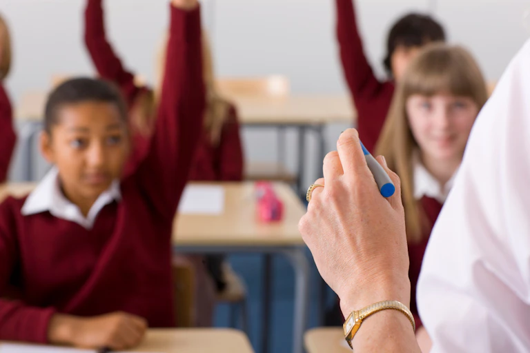 Teacher's hand in foreground, children raising hands to answer questions in background