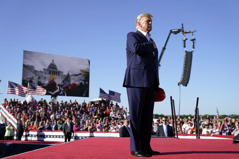 A blond older man in a blue suit stands with his hand on his heart with a video screen behind him showing an insurrection.