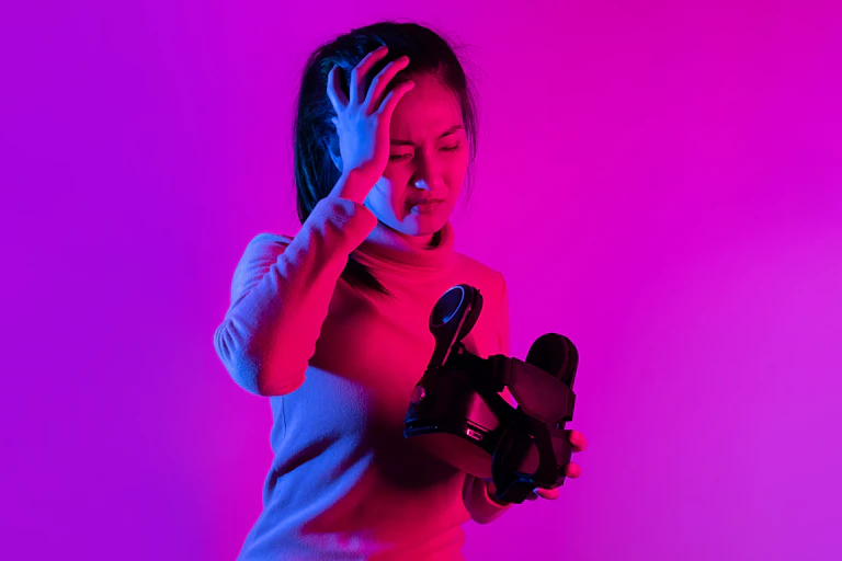 A woman holds a VR headset in one hand. Her other hand is held up to her head and her face is scrunched in pain.