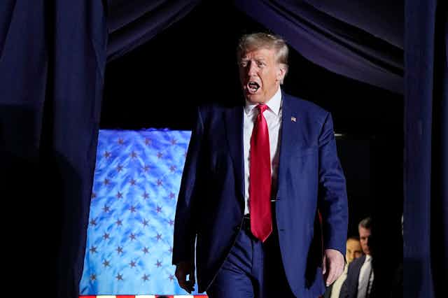 A aging blond man in a blue suit and red tie appears to be shouting as he walks onto a stage.