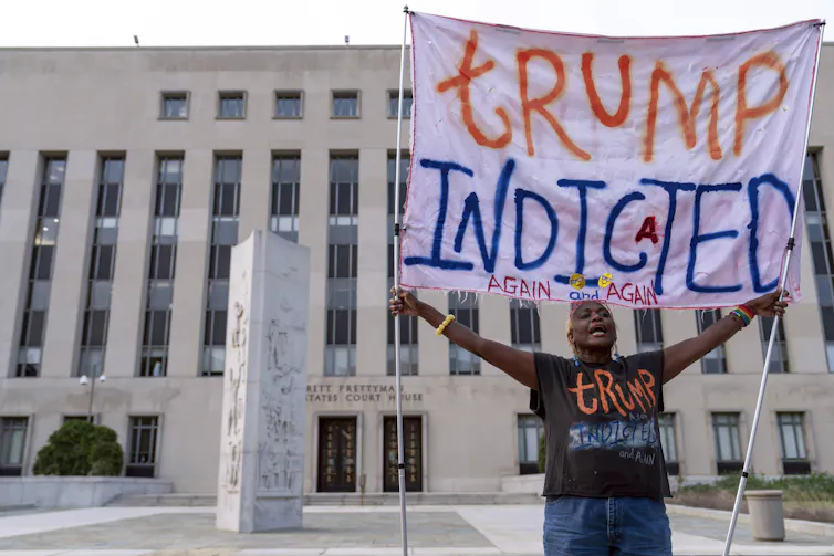 A woman holds a banner that says Trump Indicted Again and Again outside a stone building.