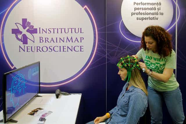 A blonde woman in a blue shirt sits as another woman with dark curly hair adjusts a green device on her head.