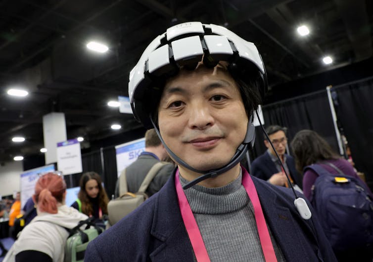 A man in a gray turtleneck stands with what looks like a black and white bike helmet on his head.