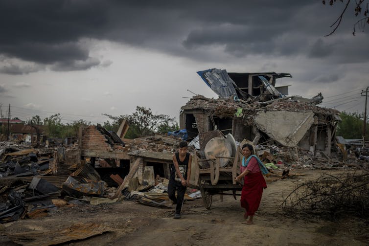 A man and woman pull a cart down a road lined with debris and damaged buildings.