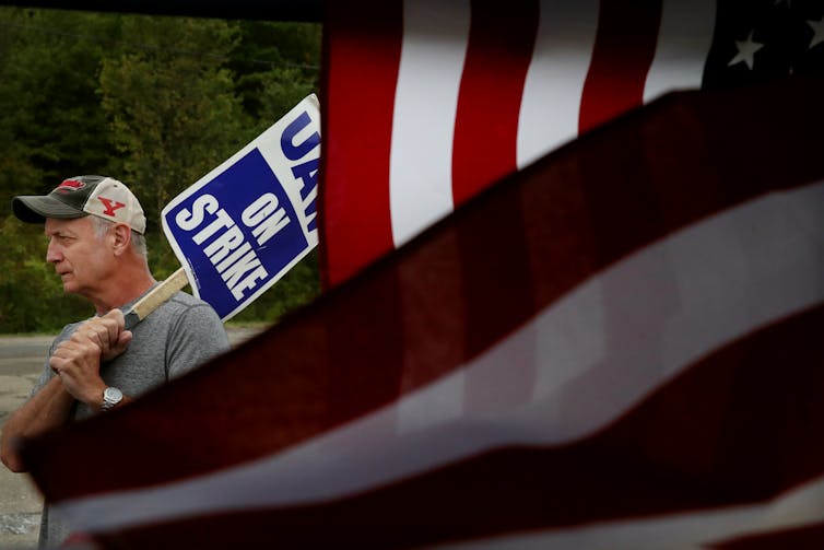 Man carries a 'UAW on strike' picket sign, enveloped in an American flag.