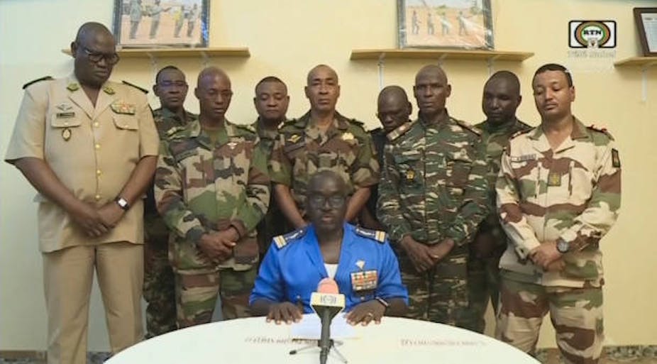Un homme assis devant un micro posé sur une table, derrière lui des hommes en tenue militaire