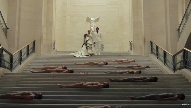 Una pareja de hombre y mujer negros descienden una escalinata coronada por una estatua sin cabeza ni brazos mientras hombres desnudos yacen en los peldaños.
