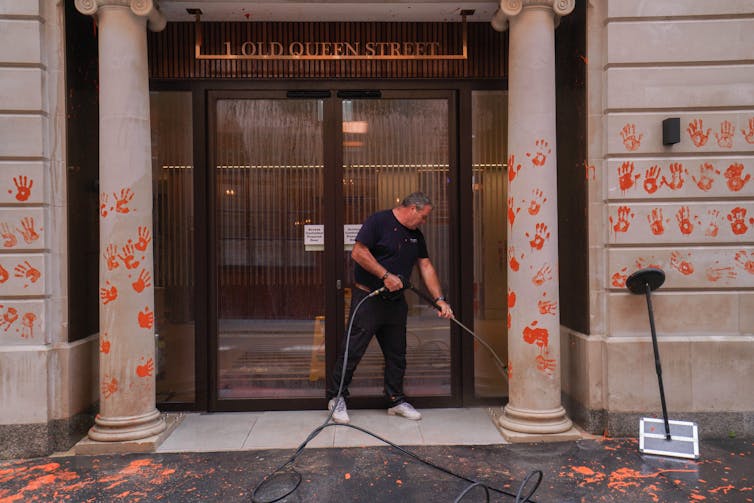 Homem limpando marcas de mãos pintadas de laranja na entrada de um prédio de escritórios.