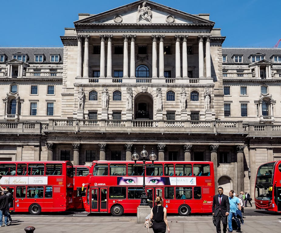 Red busses and people in front of the Bank of England building in London, blue sky.