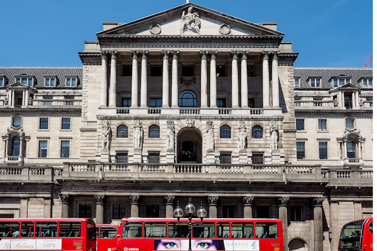 Red busses and people in front of the Bank of England building in London, blue sky.