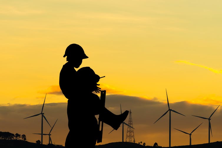 A parent with a child on their shoulders silhouetted against a wind farm at dusk.