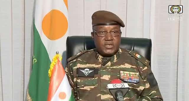Niger coup leader Gen. Abdourahmane Tchiani in military uniform flanked by the country's flag.