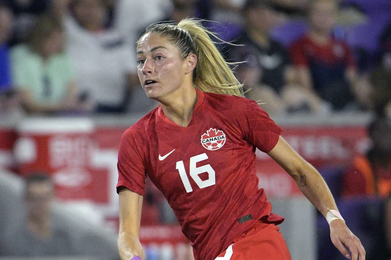 Una imagen de una mujer rubia de cintura para arriba, con una camiseta de fútbol del Equipo de Canadá y mirando hacia un campo de fútbol.