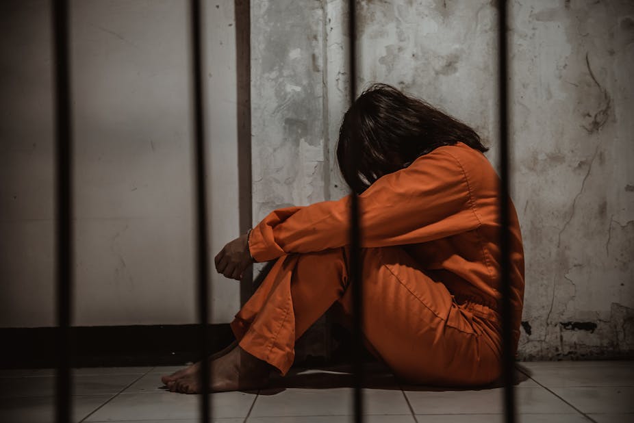 A woman in prison overalls sits behind bars with her head down.