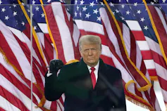 A man in a suit and tie holds up a clenched fist while he stands in front of a row of U.S. flags.