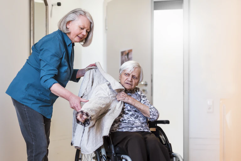 Woman taking care of old woman in wheelchair putting her jacket on.