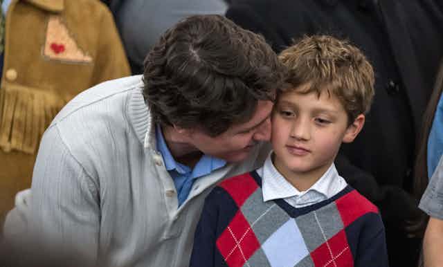 A dark-haired man whispers into the ear of a boy in an argyle sweater.