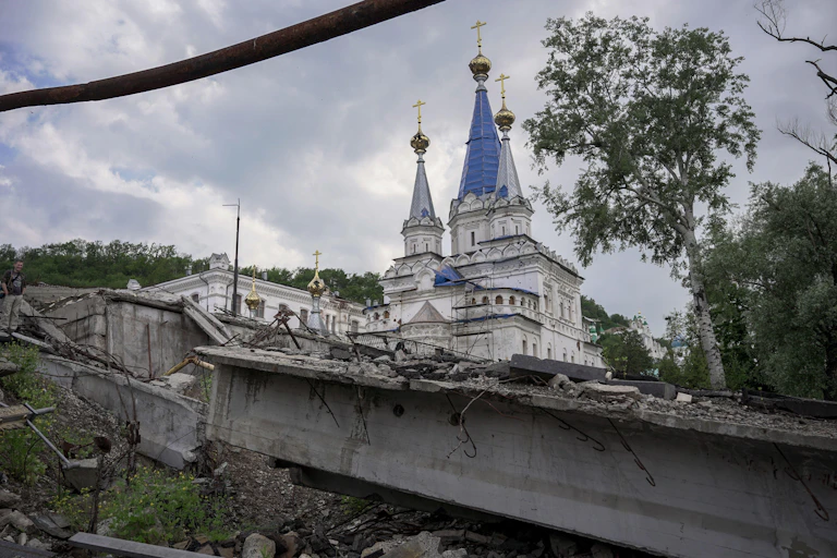 A destroyed bridge seen near a monastery, which is covered in bullet holes and fragments of shrapnel. The ruined town of Sviatohirsk in Ukraine's Donbas region.