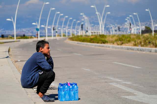 A young boy with bottles of water sits in a heat wave by the side of the road.