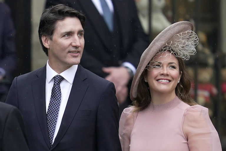Un homme aux cheveux foncés noirs porte un costume noir et une femme souriante est vêtue d'une robe et d'un chapeau roses.