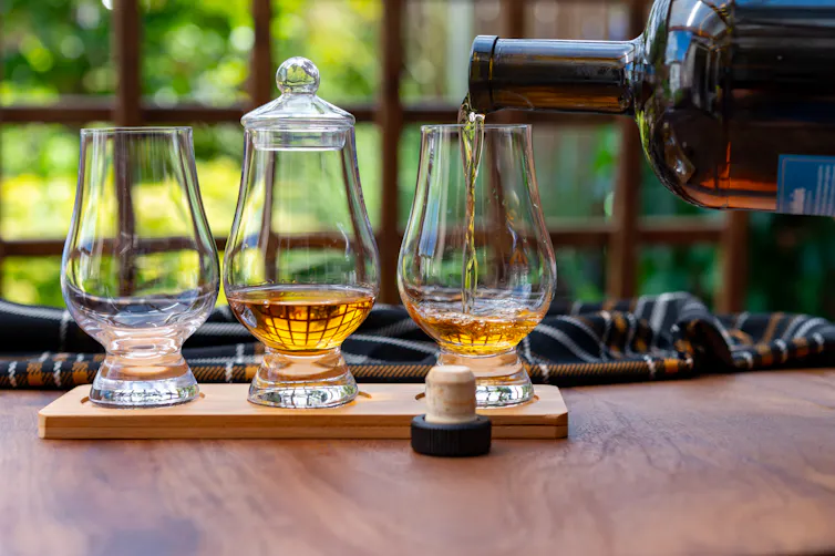 A bottle pouring brown liquid into one of three glasses, one with a glass lid, sitting on a wooden tray, all in front of a window showing greenery outside.