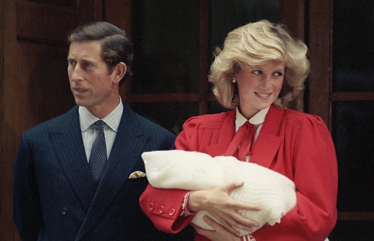 A dark-haired man looks to the side while a blond woman in red smiles while holding a swaddled baby.