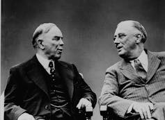 A black and white photo shows two men in suits in conversation.