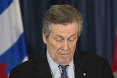 A man with brownish grey hair wearing a dark suit looks down at a news conference.